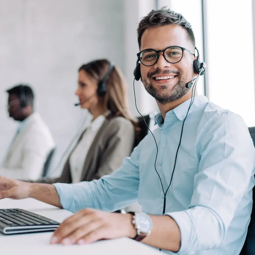 Man talar i telefon med hörlurar och har tangentbord och datorskärm framför sig i ett kontorslandskap med flera medarbetare..