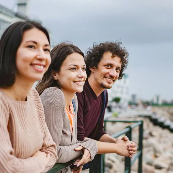 Två kvinnor och en man iklädda tröjor står vid en kaj och tittar ut över havet med bostadshus i bakgrunden.